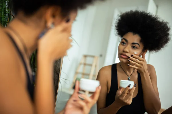 Joven mujer afroamericana aplicando crema en la cara cerca del espejo en el baño - foto de stock