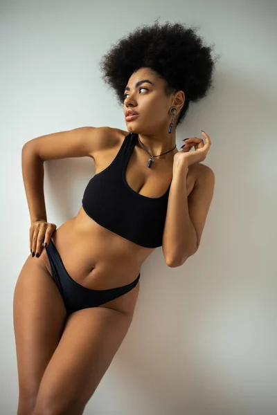 Sexy africana americana mujer en crop top y bragas tocando la cadera mientras está de pie cerca de la pared - foto de stock