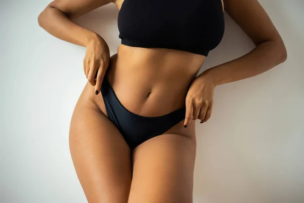 Vista ritagliata di sexy donna africana americana in crop top toccare mutandine nere vicino alla parete a casa — Foto stock