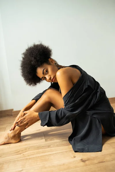 Mujer americana africana rizada con camisa tocando la pierna y mirando a la cámara mientras está sentada en el suelo - foto de stock