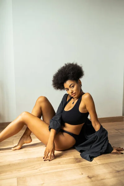 Hübsche afrikanisch-amerikanische Frau in Unterwäsche und Hemd schaut in die Kamera, während sie zu Hause auf dem Boden sitzt — Stockfoto