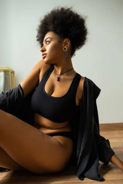 Vue latérale de la femme afro-américaine en chemise noire et sous-vêtements assis sur le sol dans la salle de bain — Photo de stock