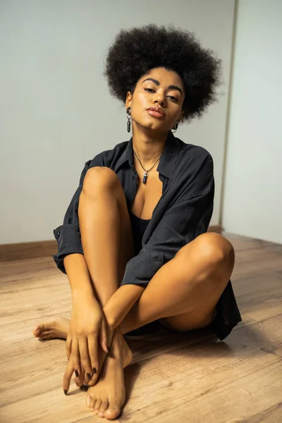 Joven mujer afroamericana en camisa negra mirando a la cámara mientras está sentada en el suelo — Stock Photo