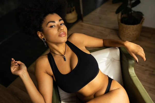 Vista ad alto angolo della donna afro-americana sexy in biancheria intima guardando la fotocamera mentre si siede sulla poltrona a casa — Foto stock