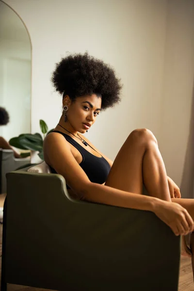 Rizado africano americano mujer en negro crop top mirando cámara mientras sentado en sillón en casa - foto de stock