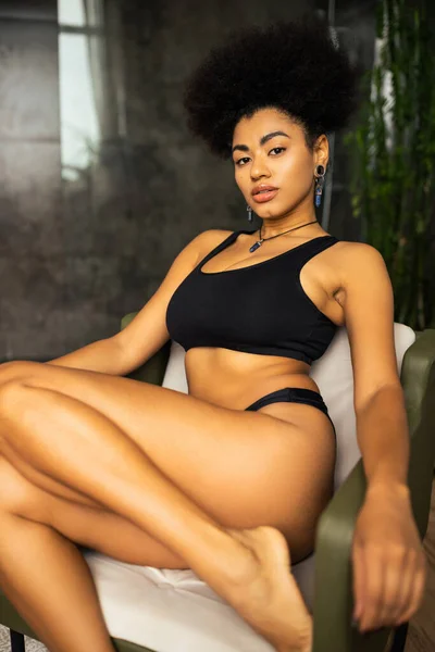 Sexy Afroamerikanerin in schwarzer Unterwäsche schaut in die Kamera, während sie zu Hause auf einem Sessel sitzt — Stockfoto