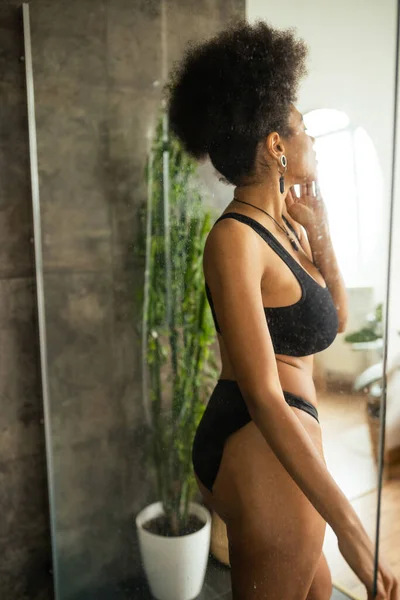 Vista laterale della giovane donna afroamericana in biancheria intima in piedi vicino alla cabina doccia a casa — Foto stock