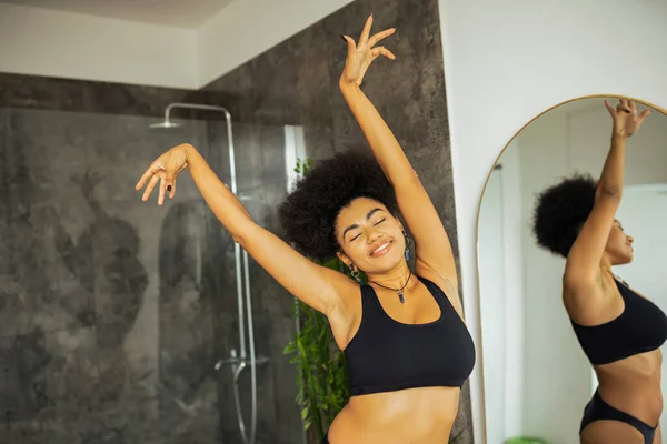 Alegre mujer afroamericana en ropa interior de pie cerca del espejo y cabina de ducha en el baño - foto de stock