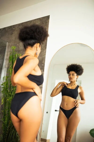 Joven mujer afroamericana tocando el cuerpo mientras mira el espejo en el baño - foto de stock