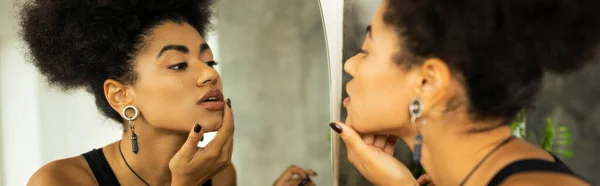 Seitenansicht einer afrikanisch-amerikanischen Frau, die ihr Kinn berührt, während sie zu Hause in den Spiegel schaut, Banner — Stockfoto