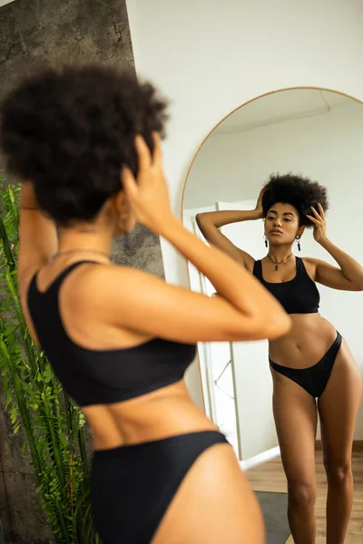 Vista posterior de la mujer afroamericana en ropa interior tocando el cabello mientras está de pie cerca del espejo en el baño - foto de stock