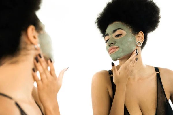 Mujer americana africana rizada con máscara de arcilla en la cara mirando el espejo en casa - foto de stock