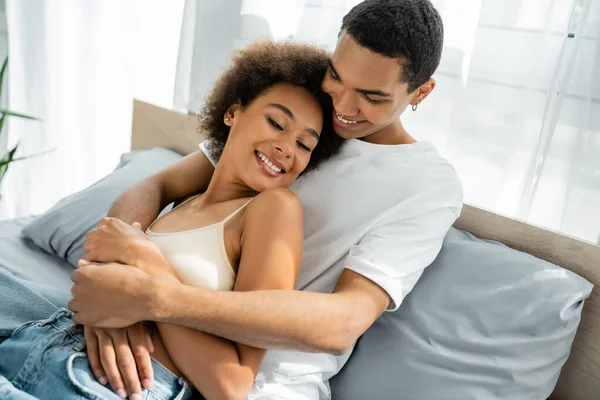 Sonriente afroamericano hombre en blanco camiseta abrazando feliz joven novia en la cama en casa — Stock Photo
