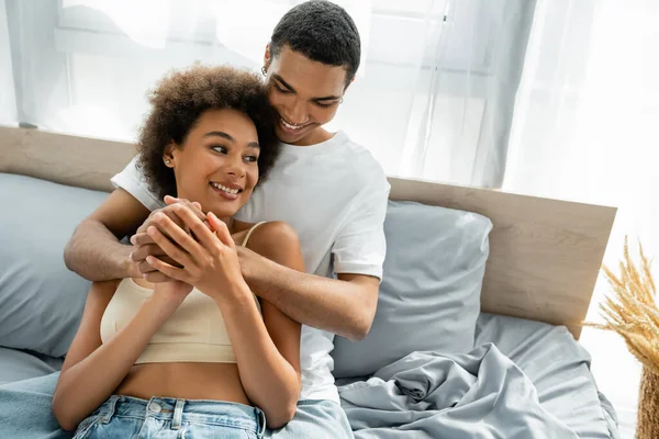 Joven africano americano pareja cogido de la mano y sonriendo en cama en casa - foto de stock