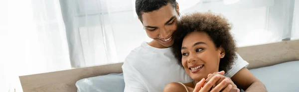 Young and curly african american woman holding hands with smiling boyfriend in bedroom, banner — Stock Photo