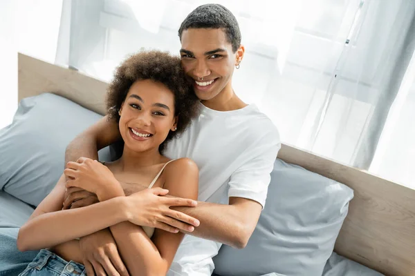 Joven afroamericano hombre con piercing abrazando feliz novia en dormitorio - foto de stock