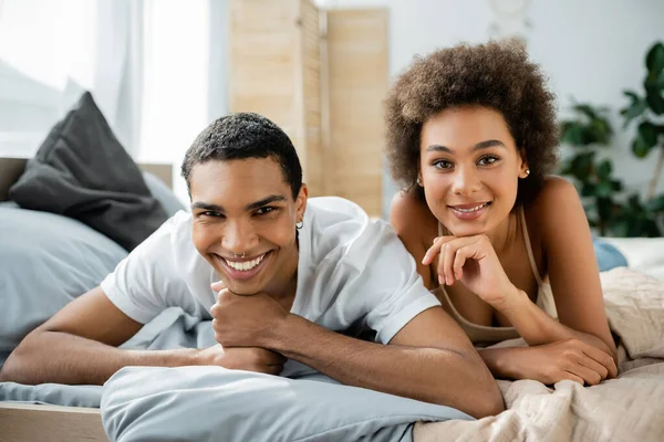 Perforado africano americano hombre con joven rizado novia acostado en la cama y sonriendo a la cámara - foto de stock
