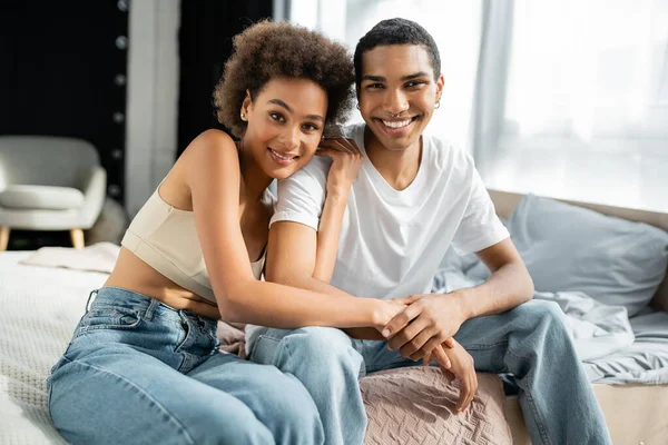 Joven y feliz afroamericano pareja en jeans sentado en la cama y mirando a la cámara - foto de stock