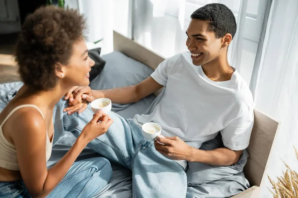 Junges afrikanisch-amerikanisches Paar mit Kaffeetassen, die einander anlächeln und Händchen halten im Schlafzimmer — Stockfoto
