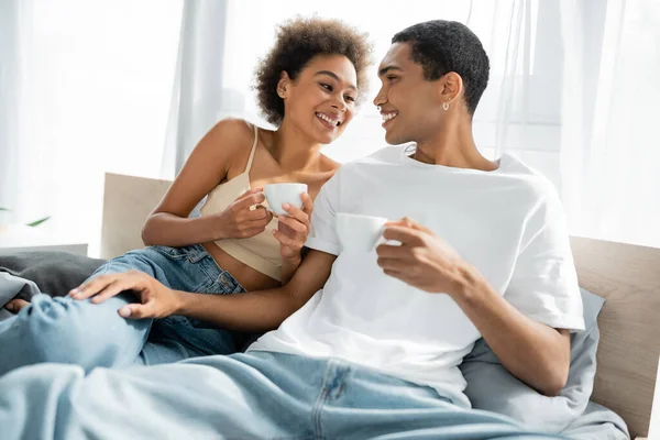 Happy african american couple with coffee cups looking at each other on bed — Stock Photo