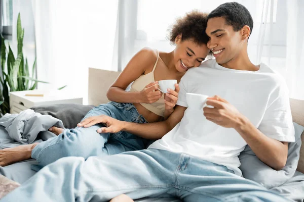 Happy african american woman with smiling boyfriend holding coffee cups on bed — Stock Photo
