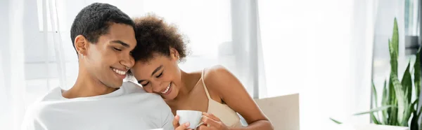 Frisé afro-américaine femme avec tasse de café souriant près du jeune petit ami dans la chambre, bannière — Photo de stock