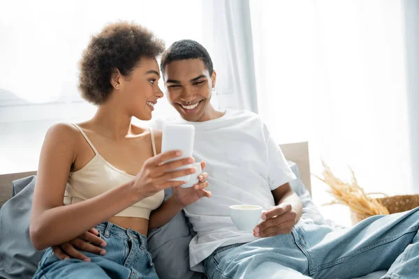 Happy african american couple with coffee cups looking at smartphone on bed — Stock Photo