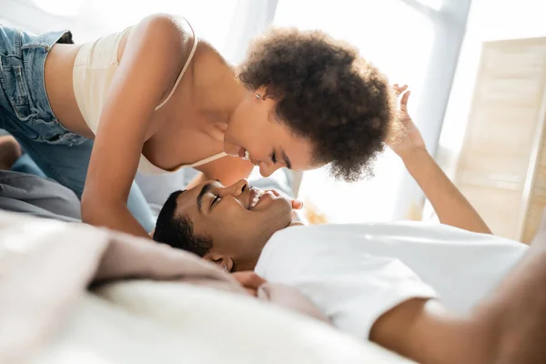 Sexy africana americana mujer sonriendo sobre novio acostado en cama en casa - foto de stock