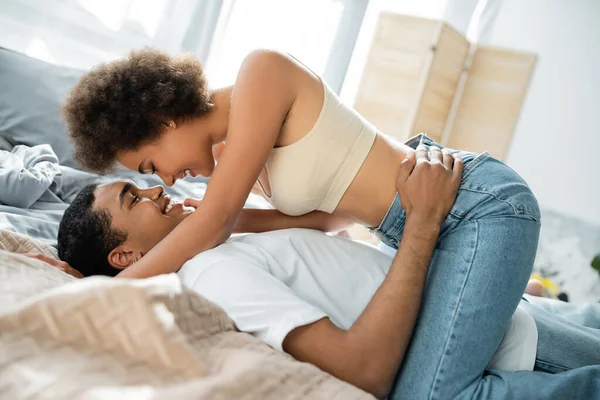 Vista lateral de joven afroamericano hombre abrazando feliz novia en jeans y top de la cosecha - foto de stock