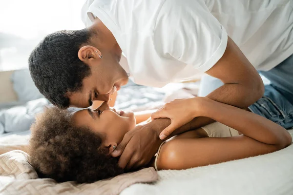 Молодой африканский американец в белой футболке обнимает счастливую девушку лежащую на кровати — стоковое фото