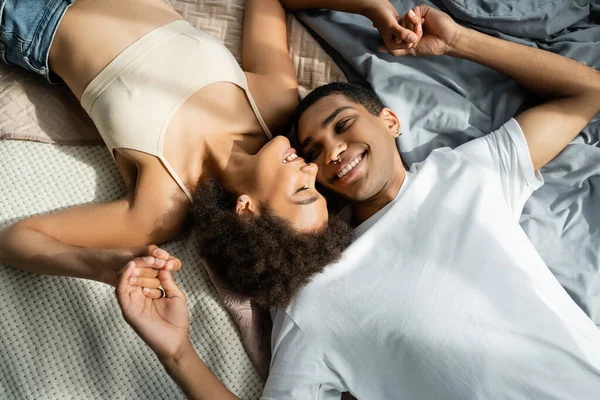 Вид сверху на молодую улыбчивую африканскую пару, лежащую на кровати и держащуюся за руки — стоковое фото