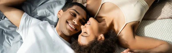 Vue du haut de sourire homme afro-américain avec piercing couché sur le lit près de petite amie heureuse, bannière — Photo de stock