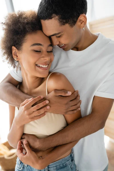 Rizado africano americano mujer sonriendo con los ojos cerrados cerca novio abrazándola en casa - foto de stock
