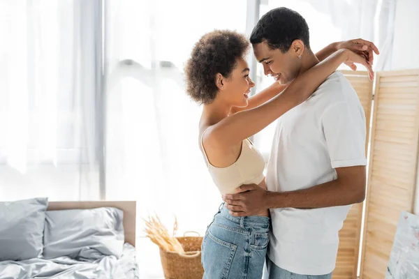 Vista lateral de la alegre pareja afroamericana abrazando mientras está de pie en el dormitorio - foto de stock