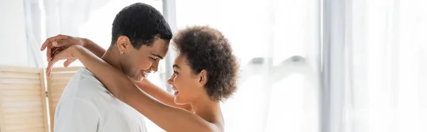 Vista lateral de la mujer afroamericana feliz abrazando novio feliz en casa, pancarta - foto de stock