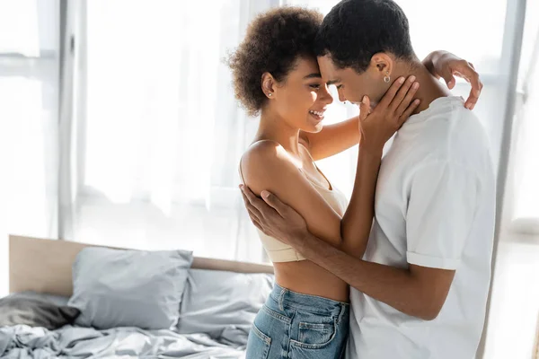 Vista lateral de los amantes afroamericanos felices abrazando cerca de la cama en casa - foto de stock