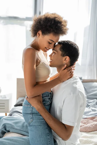 Vista lateral de la mujer afroamericana en jeans y top de la cosecha abrazando novio sonriente en casa - foto de stock