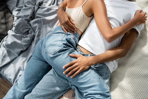 Вид сверху оборванных африканских влюбленных в джинсах, лежащих и обнимающихся на кровати — стоковое фото