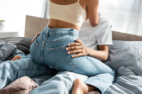 Cropped view of african american woman in jeans sitting on boyfriend on bed — Stock Photo