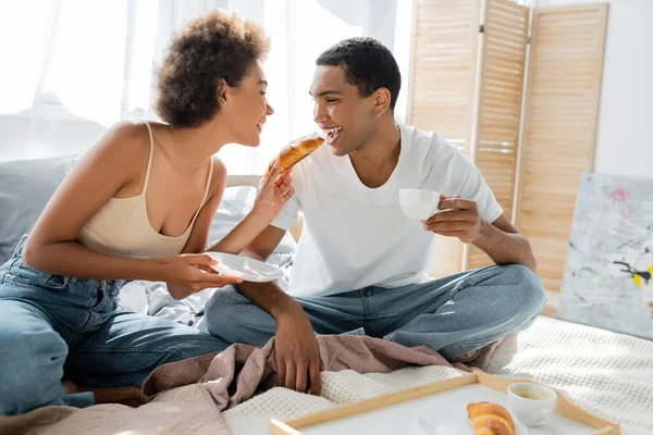 Cheerful african american woman feeding boyfriend with croissant in bedroom — Stock Photo