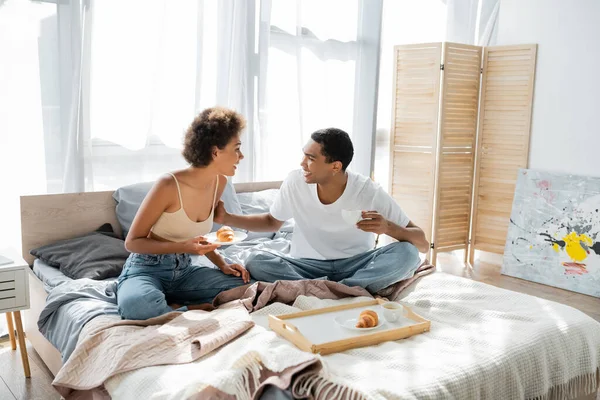 Joven afroamericano pareja con café y croissants sonriendo unos a otros en la cama - foto de stock