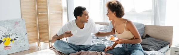 Happy african american couple with coffee and croissant talking during breakfast on bed, banner — Stock Photo