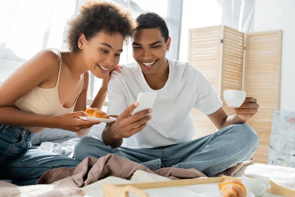 Cheerful african american couple looking at mobile phone while holding croissant in bedroom — Stock Photo