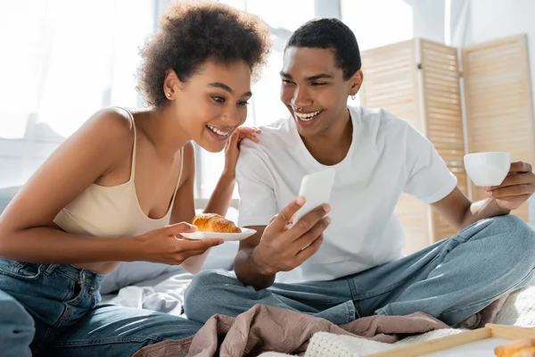 Молодий афроамериканський чоловік з чашкою кави, що показує смартфон усміхненій дівчині з круасаном у спальні — стокове фото