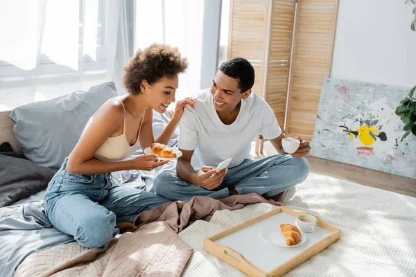Sorridente uomo africano americano in possesso di smartphone e tazza di caffè vicino fidanzata felice con croissant — Foto stock