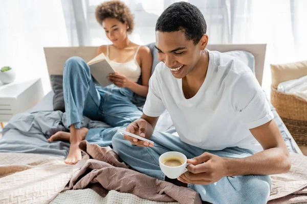 Hombre afroamericano feliz con taza de café usando teléfono inteligente cerca de novia borrosa lectura libro en la cama - foto de stock
