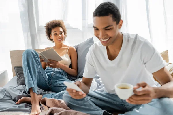 Alegre afroamericana mujer sentada en la cama con libro y mirando novio con café y teléfono móvil en primer plano borrosa - foto de stock