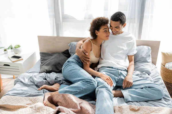 Homme afro-américain joyeux en jeans et t-shirt blanc étreignant petite amie bouclée tout en étant assis sur le lit — Photo de stock