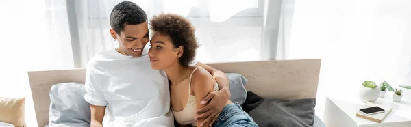 Fröhlicher afrikanisch-amerikanischer Mann im weißen T-Shirt umarmt lockige Freundin auf dem Bett, Banner — Stockfoto
