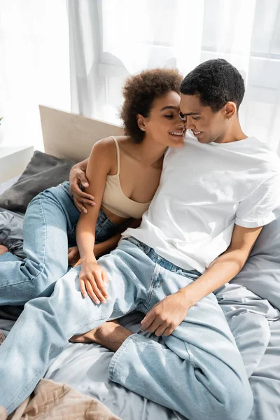 Sorridente africano americano uomo in jeans e t-shirt abbracciando giovane ragazza sul letto — Foto stock